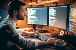 Focused young developer coding late in office writing script shown on computer monitors, serious handsome coder programmer hacker programming developing software applications working alone at night